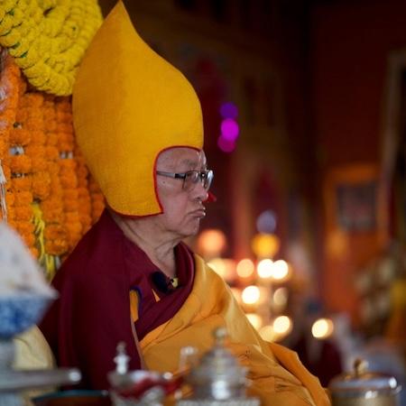 Photograph of Lama Zopa Rinpoche sitting on a teaching throne wearing a yellow pandit hat.
